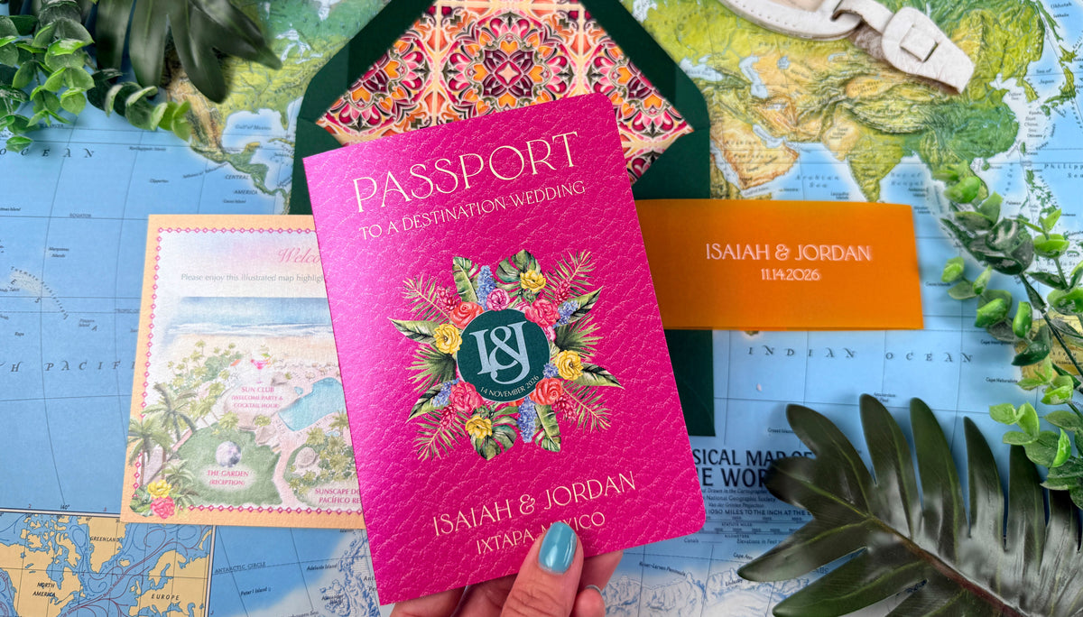 A hand holds a bright pink wedding passport-style card with floral artwork and the names Isaiah & Jordan, layered over a green patterned envelope and surrounded by travel-themed props including a map, luggage tag, and leaves.
